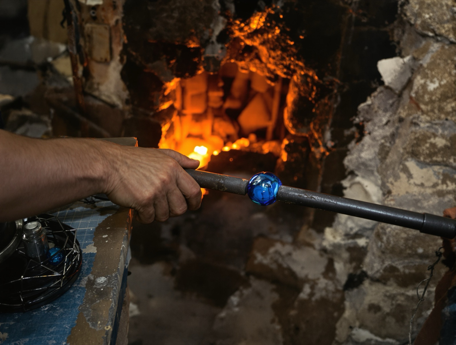 An artisan's hand turning a molten blue glass bead on a metal rod in front of a glowing kiln