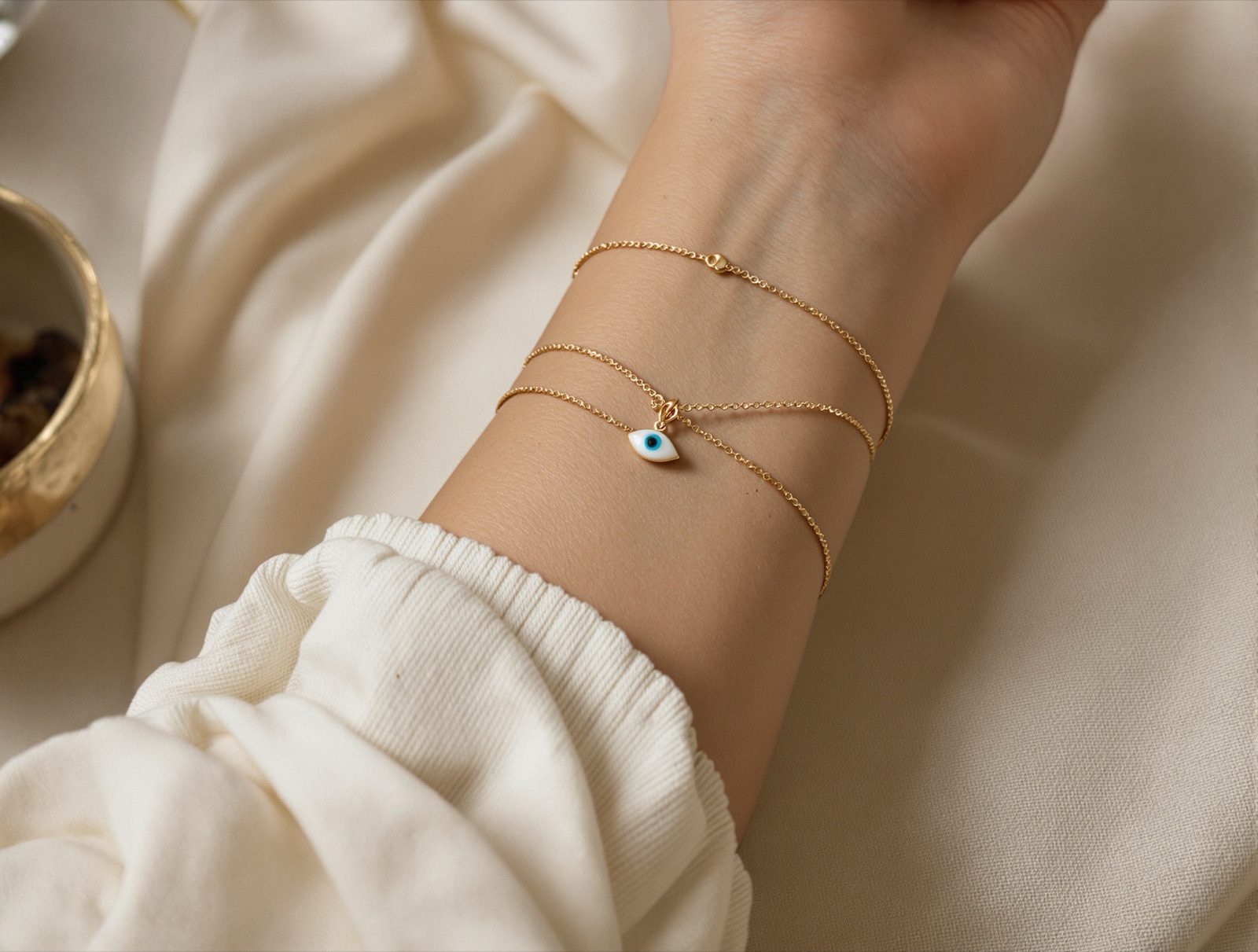A wrist wearing a fine gold chain bracelet with a small blue evil eye charm, layered with two thin bangles