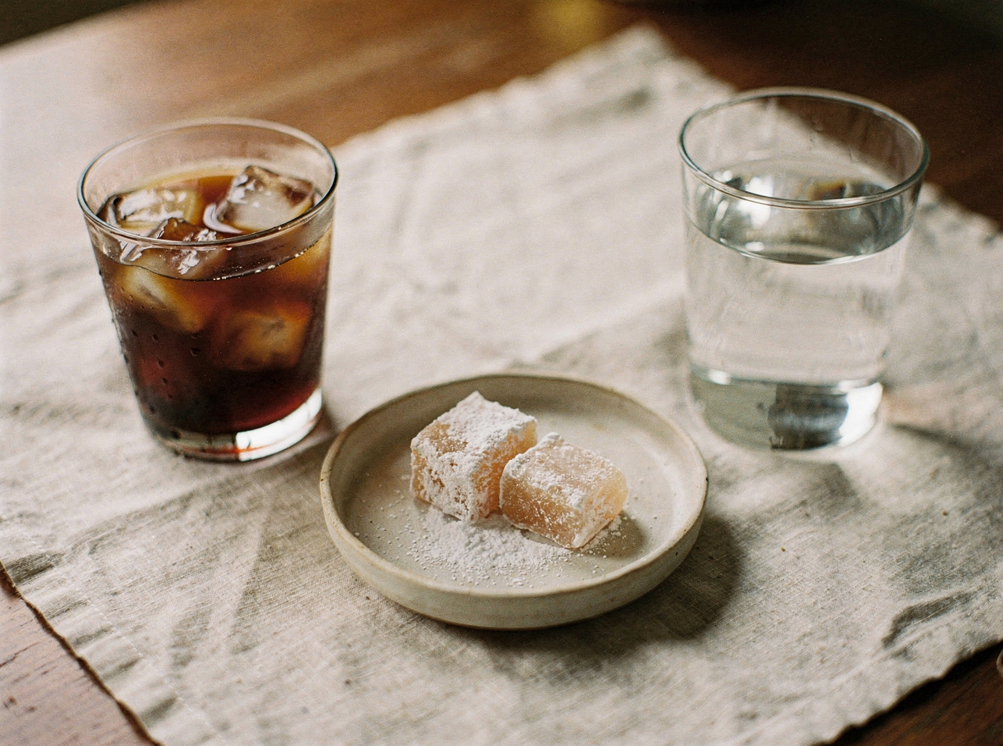 A glass of iced coffee beside a small ceramic dish of sugar-dusted lokum and a plain glass of water on a linen cloth