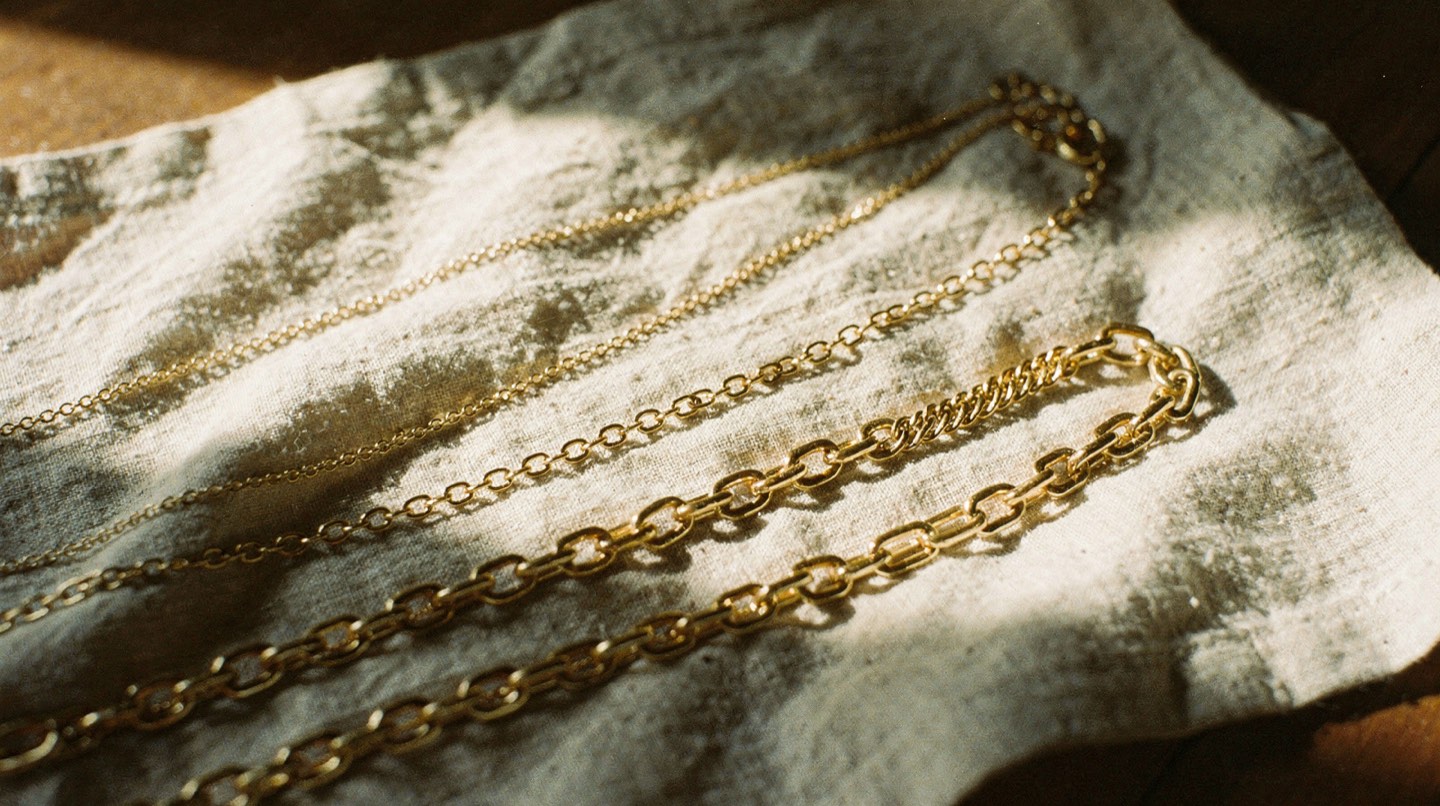 A flat lay of three gold chains of different lengths and weights arranged on a cream linen cloth