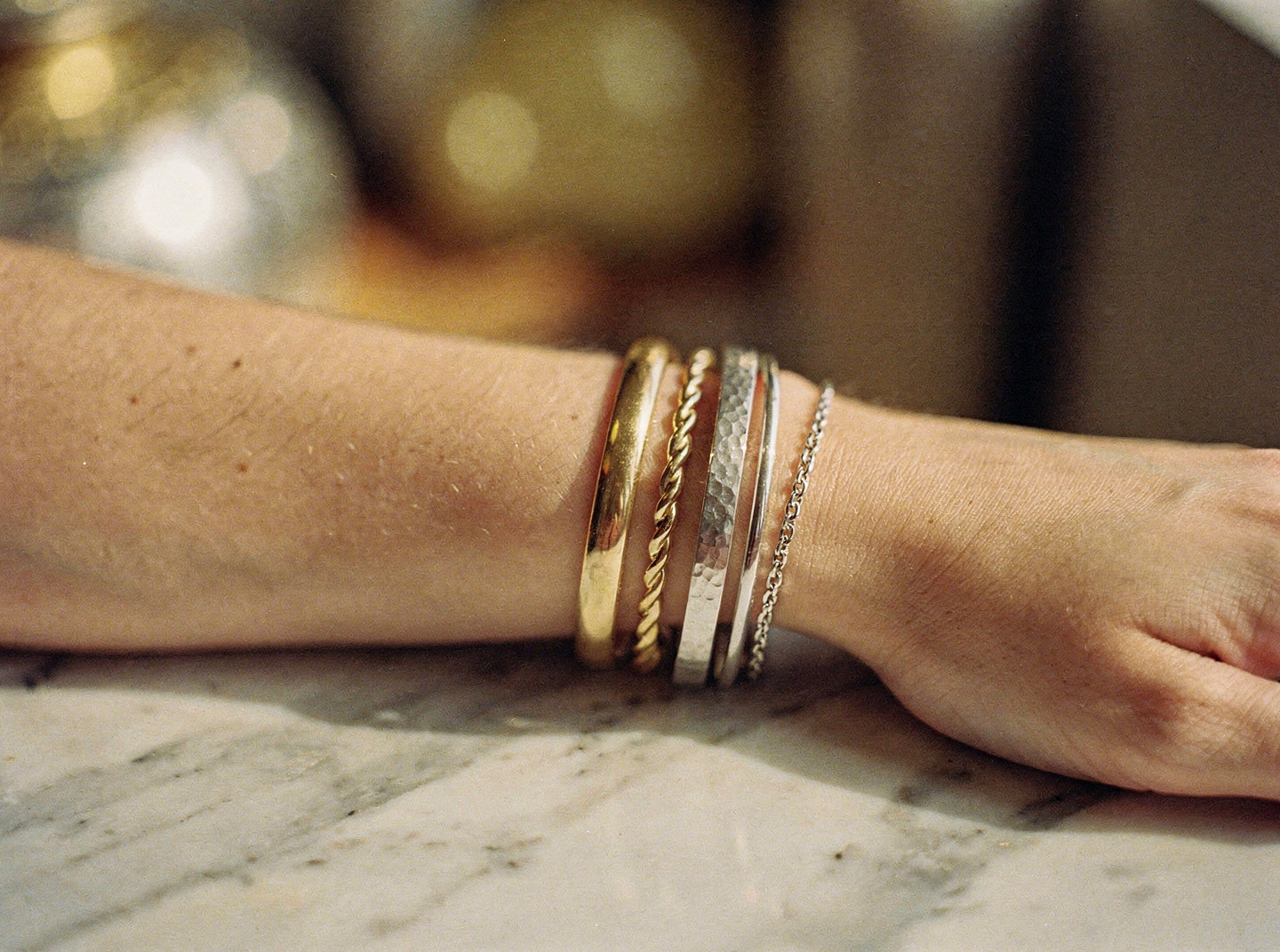 A forearm resting on a marble surface wearing a stack of mixed gold and silver bangles