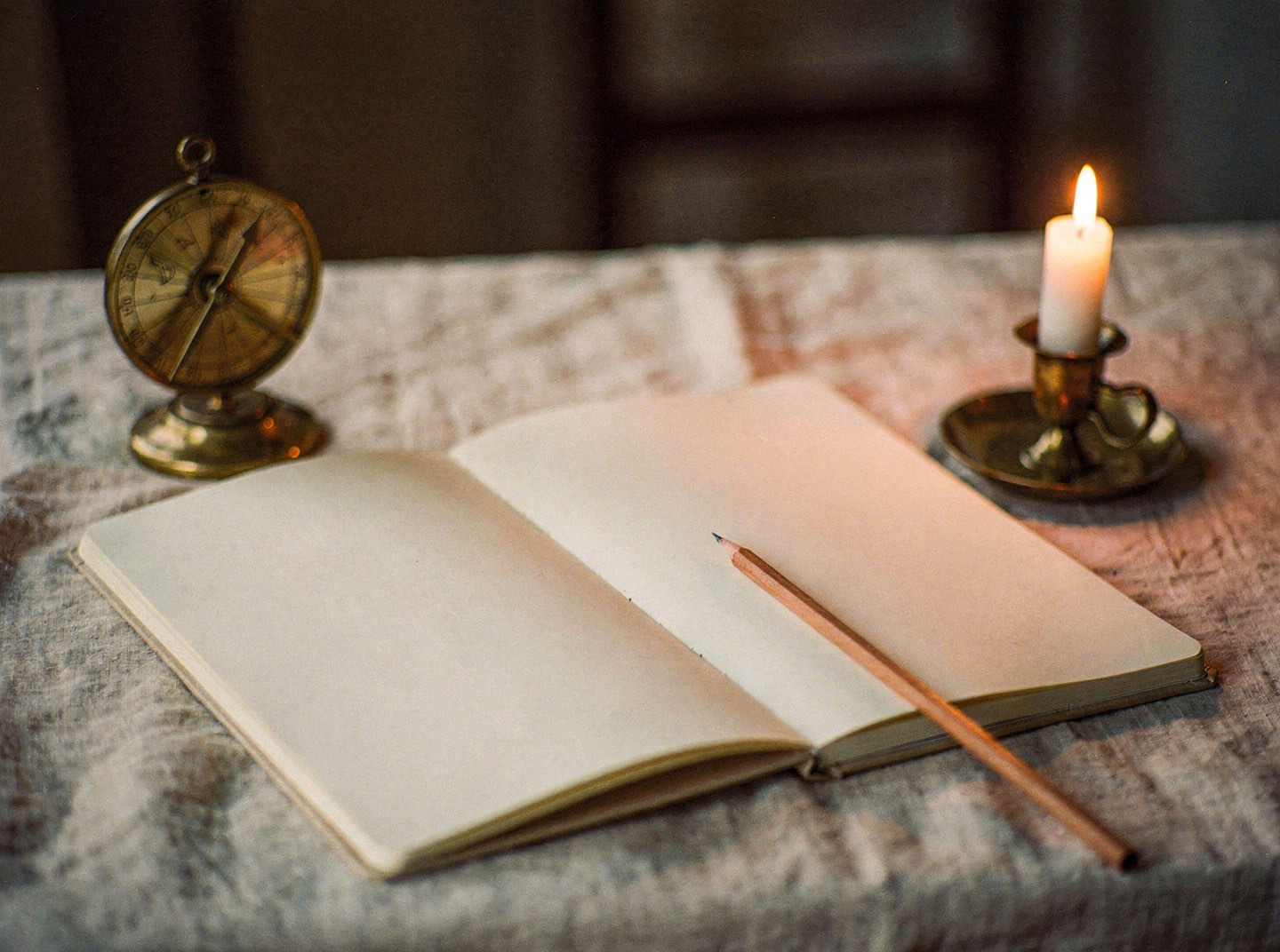 An open blank notebook and a pencil on a linen cloth beside a small brass compass and a lit candle